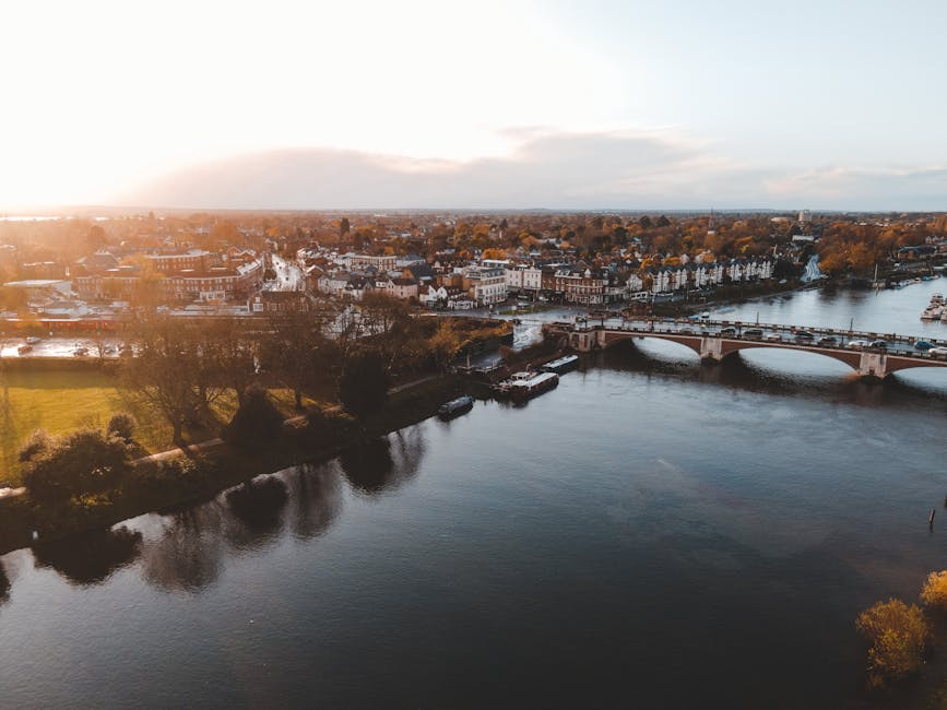 An aerial view of a riverside town in the late afternoon or early evening with the sun setting, casting a warm glow over the landscape. The river runs through the centre of the image, with several boats moored along the banks and a bridge spanning across it. On the far side of the river, there is a row of white and brick residential buildings, some with slate roofs, situated close to the water's edge. Trees with autumn foliage line the riverbanks and park areas, providing a mix of green and golden hues. The surrounding area includes open green space and pathways, indicative of a park or recreational area, with visible walking paths and benches. In the foreground, a section of a property’s doorway or entrance is visible, suggesting the image was taken from a home or property near the river. This scene may relate to house removals or property relocation services offered by Barnes Man and Van, highlighting the exterior environment connected to local moving logistics.