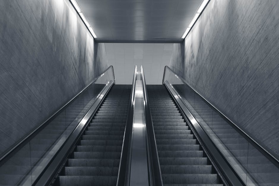 A pair of parallel escalators with black steps and silver handrails, leading upward inside a modern building with smooth gray walls and ceiling, illuminated by soft overhead lighting. The escalator on the left appears to be for ascending, while the right one is for descending, with glass side panels reflecting the surrounding environment. The clear space at the top of the escalators suggests a connection to another floor or level, which may be relevant to house removals or moving logistics in urban properties. This environment could be part of a transportation hub, such as a train station, shopping centre, or office building, where furniture and boxes from a home relocation might be transported during the moving process. Barnes Man and Van is engaged in handling complex moving challenges, including stair and access considerations for furniture transport, and such escalator environments illustrate the logistical aspects of moving large items within urban properties or commercial spaces, supporting the importance of careful planning during packing and loading processes.