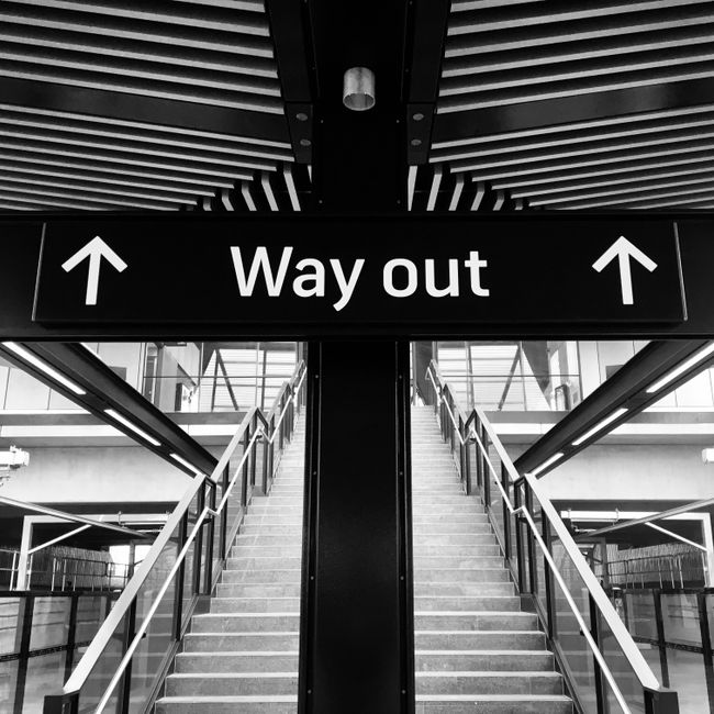 A black and white photograph of an indoor stairwell with double flight stairs leading upwards, enclosed by glass and metal railings, and illuminated by natural light from large windows at the top. Above the stairs, a rectangular black sign with white text and arrows indicates the direction for the 'Way out.' The surrounding environment appears to be part of a modern, commercial or public building, possibly an airport or train station. The image focus is on the staircase and exit signage, with no furniture or moving equipment visible, but the setting suggests it could be relevant for house removals or moving logistics, especially when considering access challenges in buildings like Barnes Station as referenced in the page title.