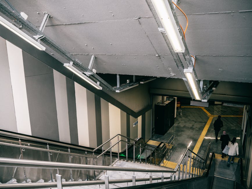 A pair of parallel escalators with black steps and silver handrails, leading upward inside a modern building with smooth gray walls and ceiling, illuminated by soft overhead lighting. The escalator on the left appears to be for ascending, while the right one is for descending, with glass side panels reflecting the surrounding environment. The clear space at the top of the escalators suggests a connection to another floor or level, which may be relevant to house removals or moving logistics in urban properties. This environment could be part of a transportation hub, such as a train station, shopping centre, or office building, where furniture and boxes from a home relocation might be transported during the moving process. Barnes Man and Van is engaged in handling complex moving challenges, including stair and access considerations for furniture transport, and such escalator environments illustrate the logistical aspects of moving large items within urban properties or commercial spaces, supporting the importance of careful planning during packing and loading processes.