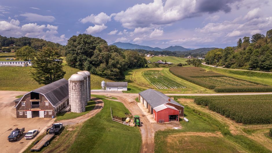 An aerial view of a rural farmstead showing a large red barn with white trim, a tall cylindrical silo attached to a nearby farmhouse, and several vehicles parked on a gravel driveway. The scene includes open fields with crops and greenery, a farmyard with various equipment, and a backdrop of forested hills under a partly cloudy sky. The image captures the outdoor environment where home relocation or furniture transport might involve loading or unloading of boxes and furniture from vehicles such as Barnes Man and Van's vans. The setting emphasizes the spacious, rural area suitable for residential moves and moving logistics in a peaceful countryside location, reflecting professional removals services across the UK.