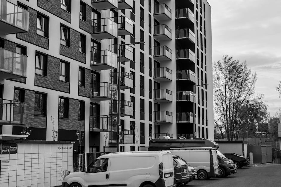 The image shows a modern multi-storey residential building with numerous balconies, some with glass railings, situated in an urban environment with trees and a cloudy sky in the background. In front of the building, there is a parking area with several vehicles, including a white van and a small cargo van, typical for home relocation and furniture transport services. The vehicles appear to be parked on a paved driveway adjacent to the building, with some equipment visible, such as a trolley or hand truck, indicating ongoing loading or unloading activities. The scene is well-lit, capturing a typical day for moving logistics involving packing and loading furniture and boxes from the property, aligning with professional removals and house moves often handled by services like Barnes Man and Van.