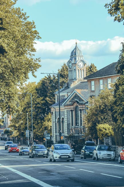 A street scene in Barnes showing several parked cars along the roadside and a few moving vehicles on the road, with a historic church featuring a prominent clock tower and a spire visible in the backg